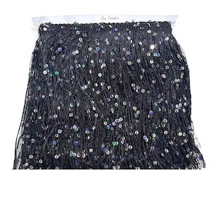 May include: Black fringe trim with iridescent sequins. The trim is densely packed with long, thin strands and small, round sequins that catch the light. The text "Sew Trends" is visible on the packaging.