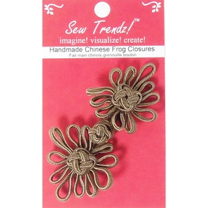 Puede incluir: Dos cierres de rana chinos hechos a mano, también conocidos como botones basculantes, de color marrón, sobre un fondo rojo. El embalaje incluye el texto "Sew Trendz!" y "Handmade Chinese Frog Closures".