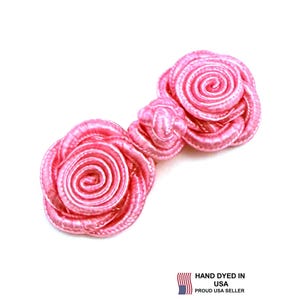 Könnte beinhalten: Ein leuchtend rosa Stoff-Knebelverschluss mit zwei rosenförmigen Knoten, die durch eine mittlere Schlaufe verbunden sind. Der Verschluss besteht aus gewickelter, satinartiger Kordel, was eine dreidimensionale florale Optik erzeugt. Der Text lautet HAND DYED IN USA PROUD USA SELLER.