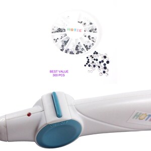 May include: A white handheld hotfix applicator tool with a blue button and a metal tip, accompanied by a circular plastic container holding clear and black rhinestones, with the text HOTEE and BEST VALUE 300 PCS visible on the items.