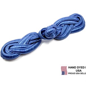 May include: A blue Chinese frog closure button featuring a woven cord design with a central knot and two symmetrical loops on either side. The button has a smooth, shiny texture and is accompanied by text that reads HAND DYED IN USA PROUD USA SELLER.