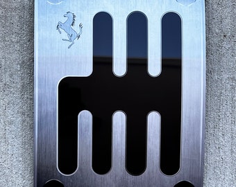 May include: A brushed metal gear shift plate with a black shift pattern. The plate is secured with four screws and features the Ferrari logo, a prancing horse, in the upper left corner. The background is a textured gray surface.