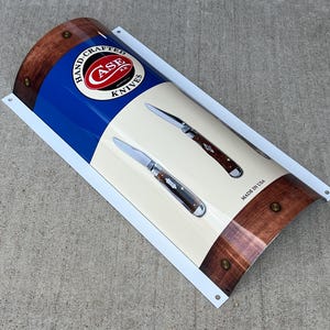 May include: A white and blue metal sign with a red Case Knives logo and two pocket knives. The sign reads "Hand-Crafted Case Knives" and "Made in USA".