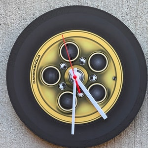 Puede incluir: Un reloj negro con forma de neumático con un borde dorado y cinco círculos negros. La esfera del reloj tiene el texto "Pirelli Cinturato P7" y "Campagnolo".