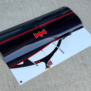 K&ouml;nnte beinhalten: Ein schwarz-wei&szlig;es Banner mit einem roten Fledermaus-Symbol und einem roten Streifen. Das Banner ist gebogen und hat einen wei&szlig;en Hintergrund mit einem schwarz-wei&szlig;en Bild eines Autos.
