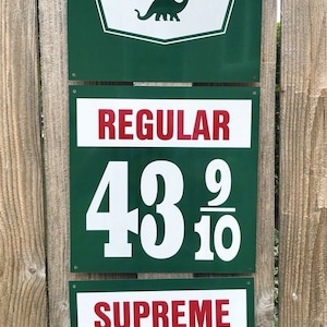 Könnte beinhalten: Ein Vintage-Schild einer Sinclair-Tankstelle mit grünem Hintergrund und weißen Buchstaben. Das Schild zeigt ein rotes Dinosaurier-Logo und den Text "Sinclair". Unter dem Logo befinden sich zwei Schilder mit weißen Zahlen auf grünem Hintergrund. Das obere Schild zeigt "REGULAR 43 9/10" und das untere Schild zeigt "SUPREME 49 9/10".