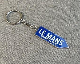 Puede incluir: Un llavero de plata con un letrero de esmalte azul que dice "LE MANS CIRCUIT des 24 H."