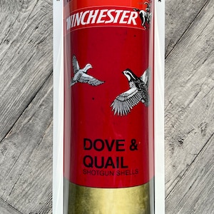 Pode incluir: Uma caixa de munição de espingarda Winchester vermelha e dourada com fundo branco. A caixa tem uma ilustração em preto e branco de uma pomba e uma codorniz em voo. O texto "Dove & Quail Shotgun Shells" está impresso na caixa.