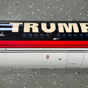 Puede incluir: Un modelo de avión blanco, rojo y negro con la palabra "TRUMP" impresa en letras blancas grandes en el lateral.