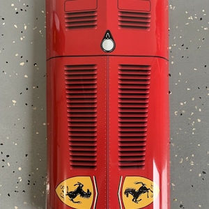 May include: A red car hood with black grill and two yellow and black horse logos that say "S.F." on either side. The hood has a white light in the center and a small Alfa Romeo logo at the bottom.