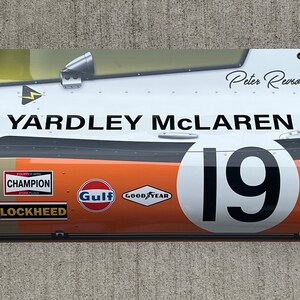 May include: A white and orange replica of a race car with the words "YARDLEY MCLAREN" and the number "19" on the side. The car has sponsors including "Champion", "Gulf", and "Goodyear". The car is signed by "Peter Revson".