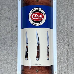 May include: A white sign with a blue and brown background featuring the Case Knives logo and three pocket knives. The sign reads "Hand-Crafted Case Knives Made in USA".