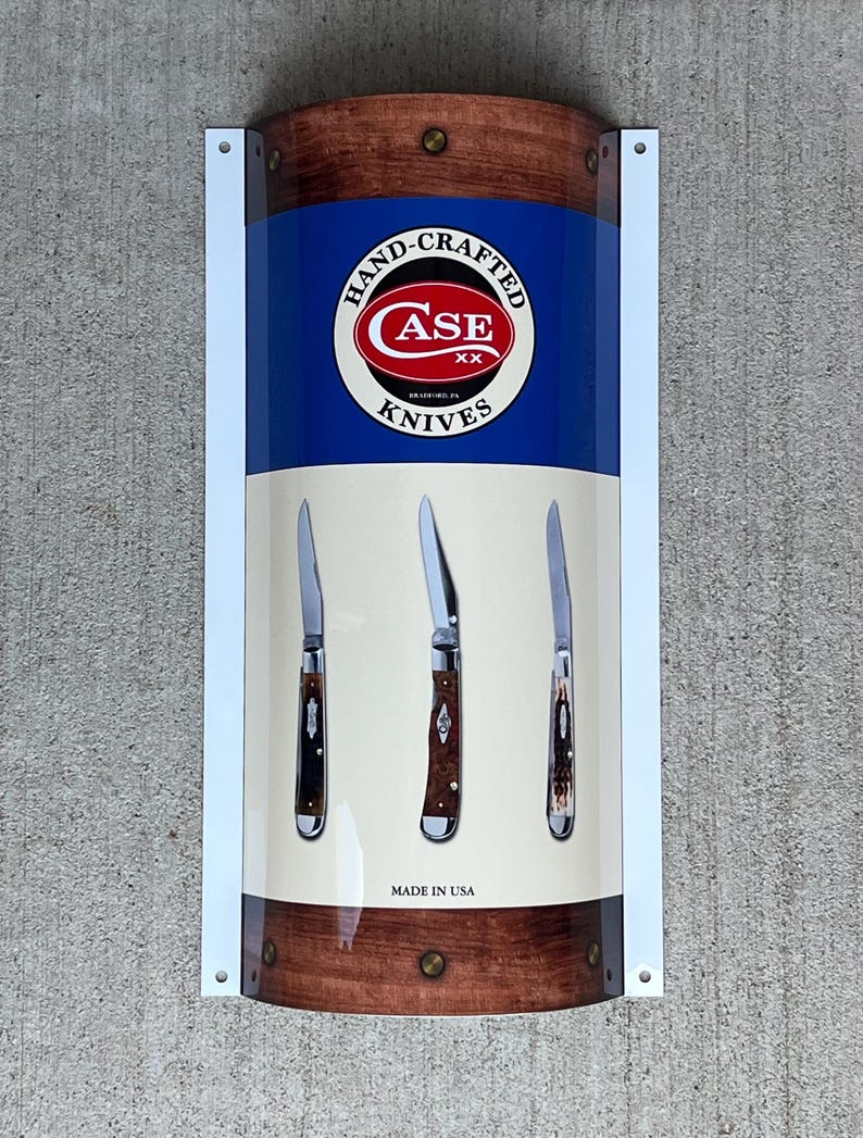 May include: A white sign with a blue and brown background featuring the Case Knives logo and three pocket knives. The sign reads "Hand-Crafted Case Knives Made in USA".