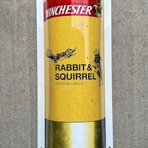 May include: A vintage-style sign featuring a yellow and gold shotgun shell design. The sign has the Winchester logo in red and white, with the text "Rabbit & Squirrel Shotgun Shells" and illustrations of a rabbit and squirrel.