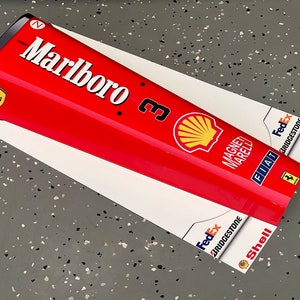 May include: A red, white, and black Formula One race car nose cone with the Marlboro logo, Shell logo, and other sponsors. The nose cone is on a gray surface.