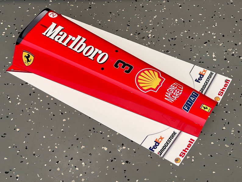 May include: A red, white, and black Formula One race car nose cone with the Marlboro logo, Shell logo, and other sponsors. The nose cone is on a gray surface.
