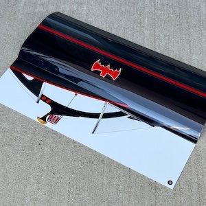 K&ouml;nnte beinhalten: Ein schwarzes und rotes Banner mit wei&szlig;em Hintergrund, das eine stilisierte Illustration des Batmobils aus der Batman-Fernsehserie der 1960er Jahre zeigt. Das Batmobil ist aus einem niedrigen Winkel dargestellt, wobei die Front des Autos zum Betrachter zeigt. Das Batmobil hat einen roten Streifen, der entlang der Seite verl&auml;uft, und ein rotes Fledermaussymbol auf der Motorhaube. Das Banner hat &Ouml;sen in den Ecken.