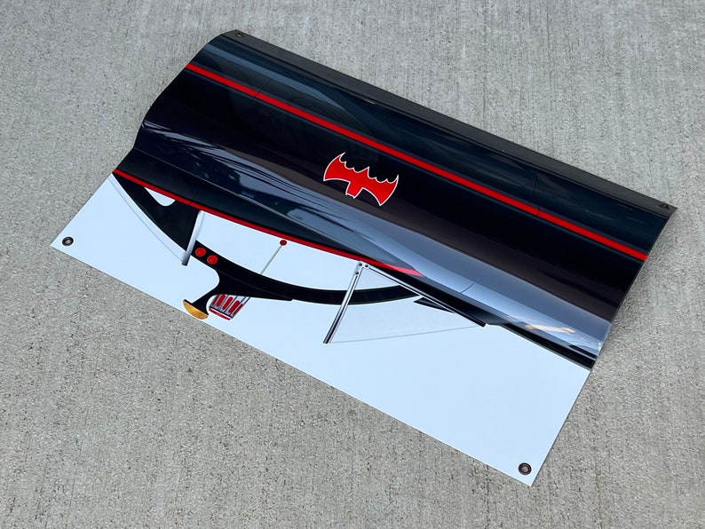 K&ouml;nnte beinhalten: Ein schwarzes und rotes Banner mit wei&szlig;em Hintergrund, das eine stilisierte Illustration des Batmobils aus der Batman-Fernsehserie der 1960er Jahre zeigt. Das Batmobil ist aus einem niedrigen Winkel dargestellt, wobei die Front des Autos zum Betrachter zeigt. Das Batmobil hat einen roten Streifen, der entlang der Seite verl&auml;uft, und ein rotes Fledermaussymbol auf der Motorhaube. Das Banner hat &Ouml;sen in den Ecken.