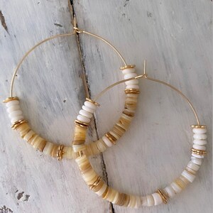 May include: A pair of gold hoop earrings with white and beige shell beads. The earrings are large and have a simple, elegant design.