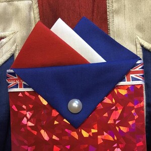 May include: A red, white, and blue fabric pocket square with a pearl button. The pocket square is folded into a triangle shape and is sitting on a red, iridescent fabric background.