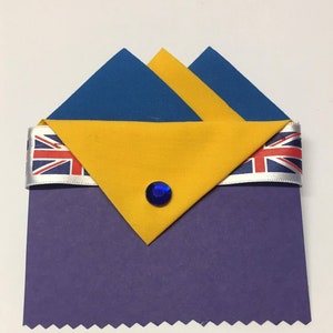 May include: A purple fabric pocket square with a yellow and blue fabric pocket square folded on top. The pocket square is decorated with a blue jewel and a white ribbon with a Union Jack pattern.