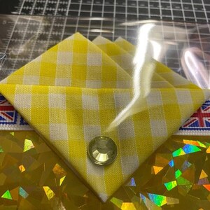 May include: A yellow and white gingham fabric with a clear plastic overlay. A single rhinestone embellishment is centered on the fabric.