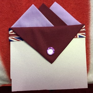 May include: A white envelope with a burgundy fabric flap and a purple button. The flap is decorated with a ribbon featuring a Union Jack pattern.