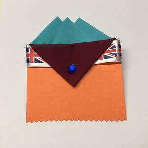 May include: A folded teal fabric square with a maroon fabric flap, topped with a white ribbon with a Union Jack pattern. A blue jewel is centered on the flap. The fabric is placed on a bright orange card with a scalloped edge.