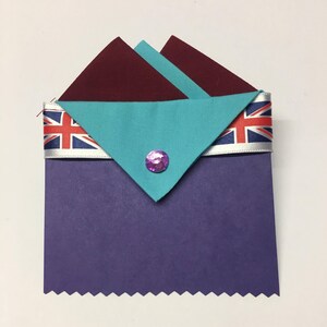 May include: A purple card with a scalloped edge, decorated with a turquoise and maroon fabric triangle with a pink gem in the center. The triangle is topped with a white ribbon with a red, white, and blue Union Jack design.
