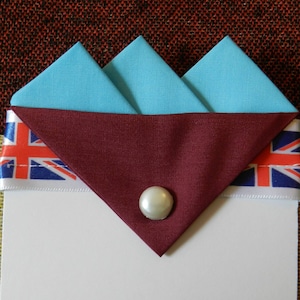 May include: A folded blue fabric pocket square with a maroon fabric trim and a white pearl button. The pocket square is sitting on top of a white card with a red, white, and blue ribbon with a Union Jack design.