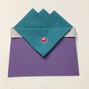 May include: A teal fabric envelope with a pink button closure, folded over a purple cardstock base with a white ribbon.