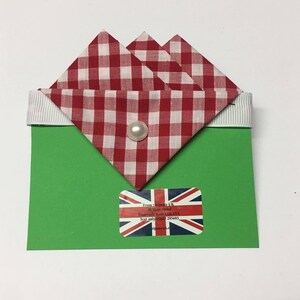 May include: A green card with a red and white gingham fabric bow on top. The bow is folded into a triangle shape and has a white pearl in the center. The card has a sticker with the Union Jack flag and text that reads "From: Hipster UK, 38 West Street, Dunstable Beds LU6 1TA, Text only 1800 293693, hipsteruk.co.uk".