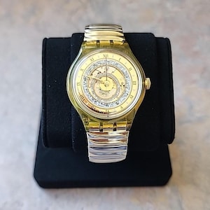 May include: A gold-toned Swatch watch with a transparent yellow case and a silver-tone band. The watch face features Roman numerals and intricate detailing. The watch is displayed on a black stand.