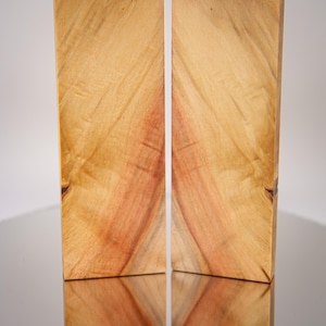 May include: Two rectangular wooden blocks with a natural wood grain pattern. The wood has a warm, light brown color with reddish-brown streaks. The blocks are standing upright, and their reflection is visible below.