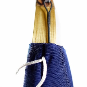 Puede incluir: Un mango de madera con veta marr&oacute;n claro est&aacute; parcialmente cubierto por una funda de tela azul oscuro. Un cord&oacute;n negro fino est&aacute; sujeto a la parte superior del mango y un hilo blanco atraviesa la tela cerca de la base de la madera.
