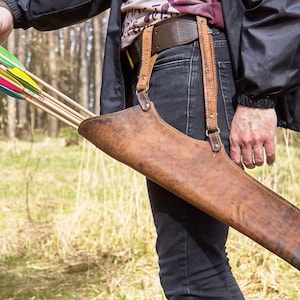 May include: A brown leather archery quiver, worn on the hip, holds several arrows with colourful fletching. The quiver is attached to a belt and braces. The background is a blurred forest scene.