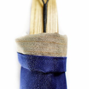 Puede incluir: Una funda de almacenamiento vertical con una base de tela texturizada azul profundo y un borde superior doblado de material tejido color canela claro. Un mango de madera sobresale por arriba, asegurado por un cord&oacute;n el&aacute;stico negro en una leng&uuml;eta de cuero marr&oacute;n.