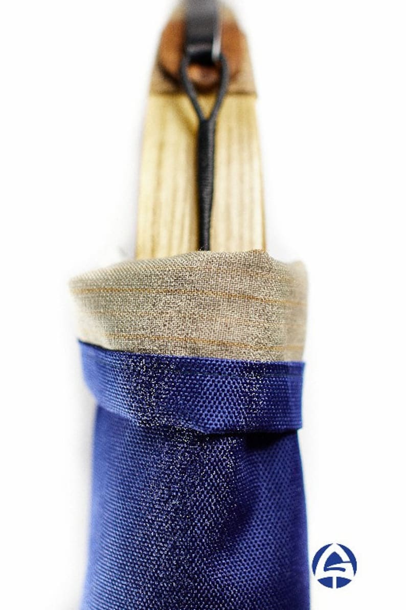Puede incluir: Una funda de almacenamiento vertical con una base de tela texturizada azul profundo y un borde superior doblado de material tejido color canela claro. Un mango de madera sobresale por arriba, asegurado por un cord&oacute;n el&aacute;stico negro en una leng&uuml;eta de cuero marr&oacute;n.