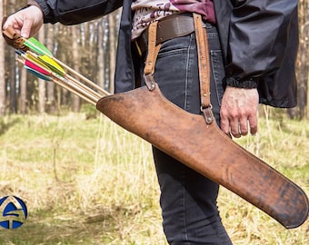 Könnte beinhalten: Ein brauner Lederköcher, der an der Hüfte getragen wird, enthält mehrere Pfeile mit farbigen Befiederungen. Der Köcher ist an einem Gürtel und Hosenträgern befestigt. Der Hintergrund ist eine verschwommene Waldszene.