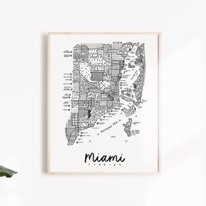 May include: Black and white illustrated map of Miami, Florida, in a wooden frame. The map details the city's neighborhoods and streets. The word "Miami" is written in cursive below the map.