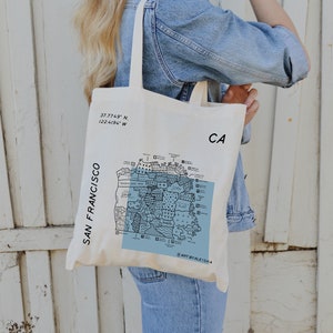 May include: A white canvas tote bag with a black and blue map of San Francisco, California. The bag has the coordinates 37.7749° N, 122.4194° W printed on the front. The text "ART BY ALEISHA" is printed on the bottom of the map.