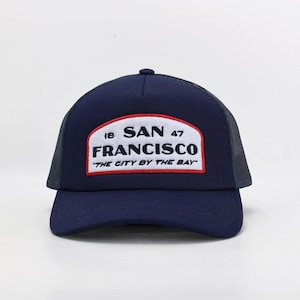 May include: Navy blue trucker hat with a mesh back and a curved brim. The front features a white patch with "SAN FRANCISCO" and "THE CITY BY THE BAY" text in navy blue and red. A classic baseball cap style.