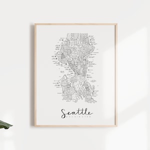 May include: A black and white line art print of a map of Seattle, Washington. The print is framed in a light wood frame and is hanging on a white wall.