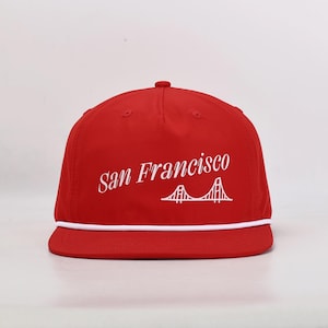 May include: A red baseball cap with a white rope detail and a flat brim. The cap features the text "San Francisco" in a script font, along with a white graphic of the Golden Gate Bridge.
