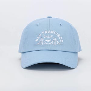 Puede incluir: Gorra de béisbol azul claro con visera curva. La gorra presenta texto bordado en blanco que dice "SAN FRANCISCO CALIF." sobre un gráfico del Golden Gate Bridge y un pájaro volando.