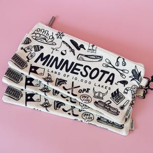 May include: Three white canvas zipper pouches with black illustrations and the word "MINNESOTA" in large letters. The pouches are decorated with drawings of state symbols and the text "LAND OF 10,000 LAKES". The background is pink.