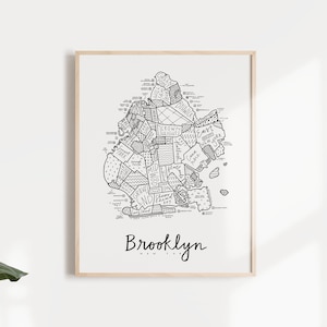 May include: A black and white line art print of a map of Brooklyn, New York. The map shows the different neighborhoods of Brooklyn with their names written in a handwritten style. The print is framed in a light wood frame and is hanging on a white wall.