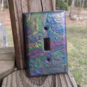 May include: A light switch cover with a colorful abstract design featuring shades of blue, green, purple, and gold. The design resembles a swirling, marbled pattern.