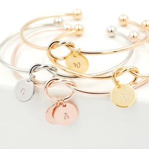 May include: Three gold, silver, and rose gold bangle bracelets with a knot charm and a personalized disc charm. The charms are engraved with the letters "W", "E", and "A".