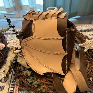 Peut inclure: Un sac en cuir marron avec un motif d'aile de dragon en cuir beige. Le sac a une bandoulière et est posé sur une couronne.